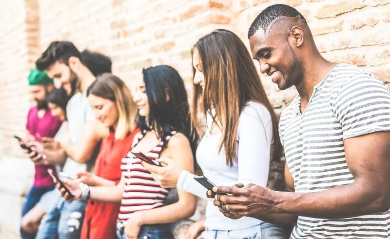 A group of diverse individuals focused on their smartphones while standing against a brick wall.