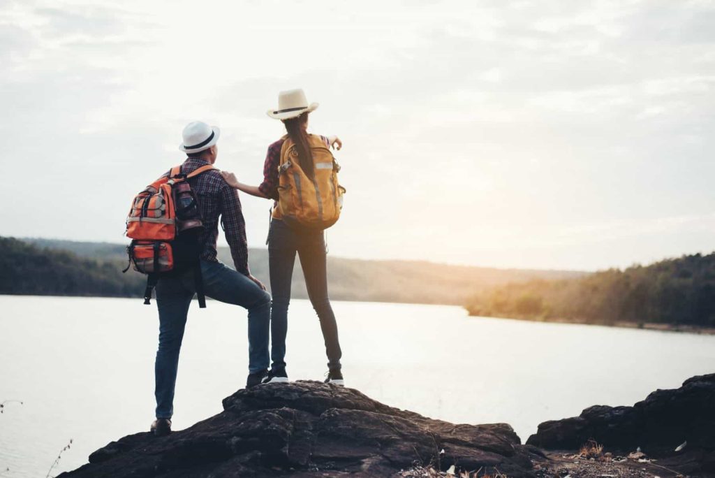 Two hikers with backpacks standing on a rock overlooking a lake at sunset.