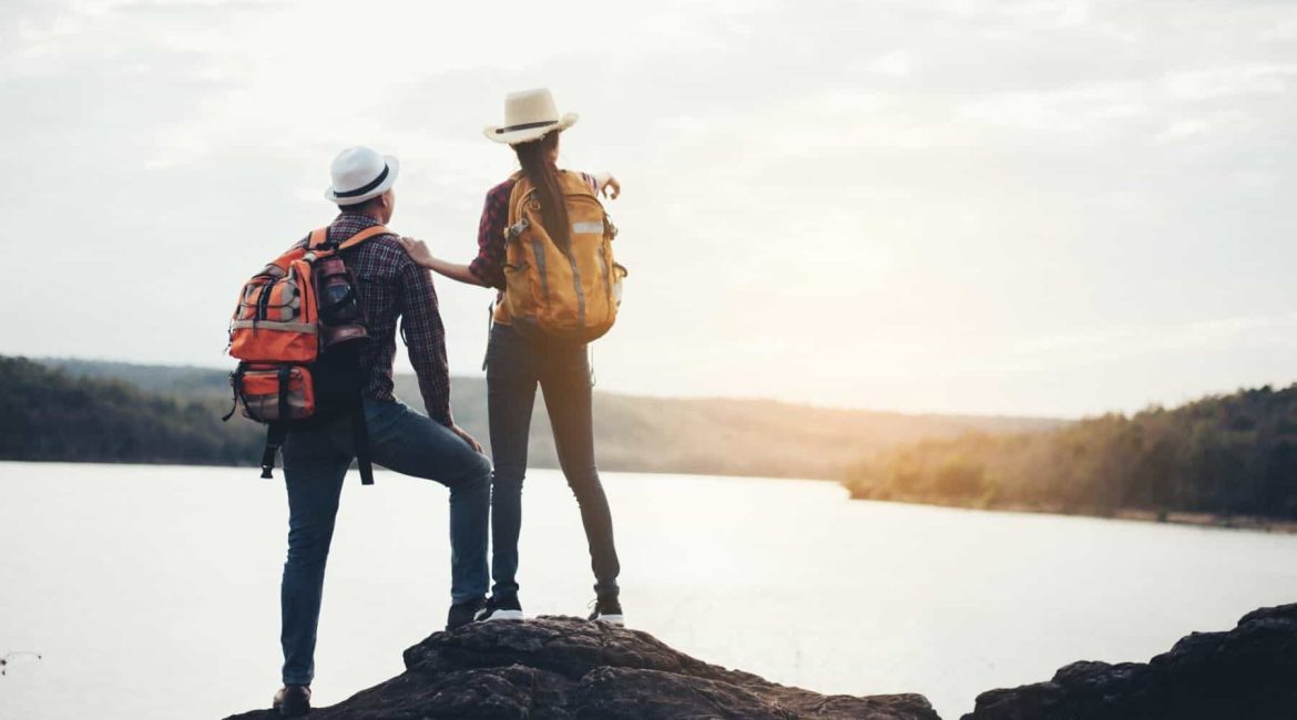 Two hikers with backpacks standing on a rock overlooking a lake at sunset.