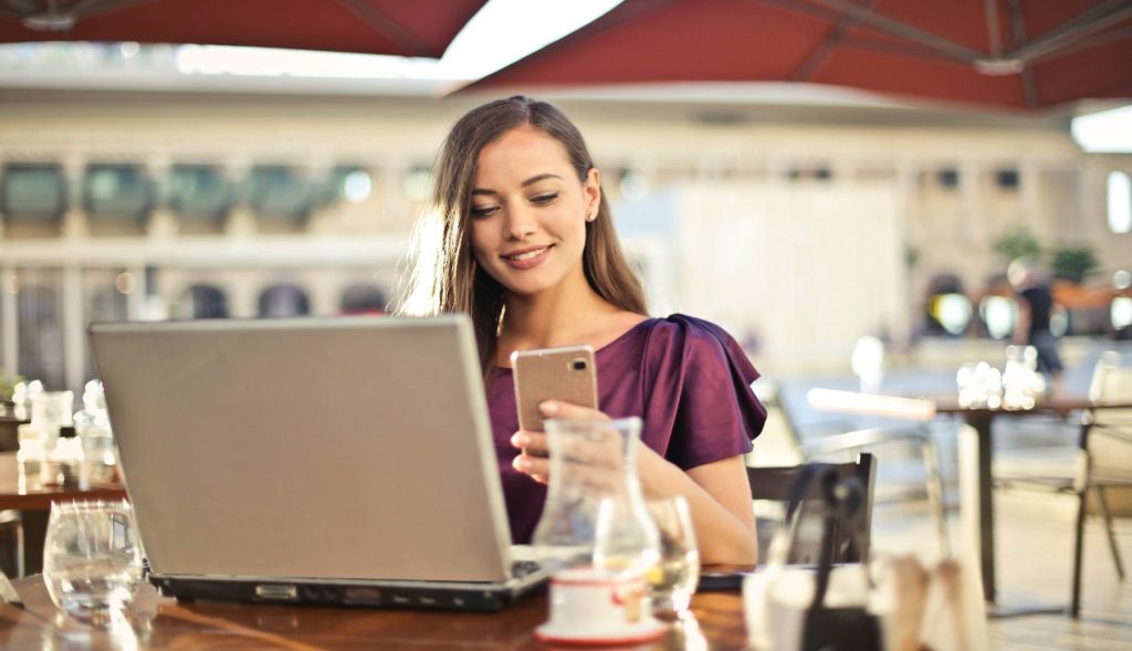 Woman working on laptop and using smartphone at an outdoor cafe, enjoying a sunny day.