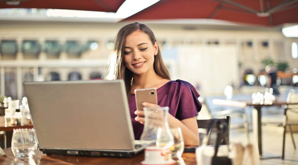Woman working on laptop and using smartphone at an outdoor cafe, enjoying a sunny day.