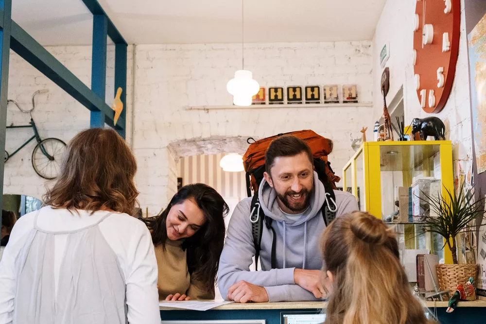 Two people conversing with a receptionist in a cozy, eclectic hostel lobby.