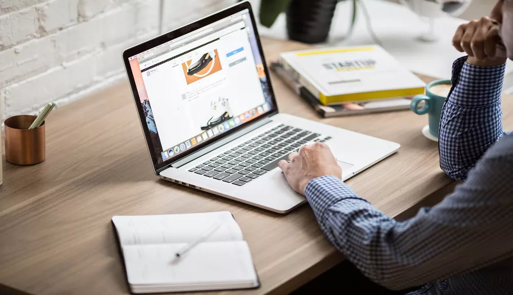 AAMP Agency - 5 Ways to Boost the Credibility of Your Adventure Tourism Website 2 Person using laptop on desk with coffee mug and notepad.