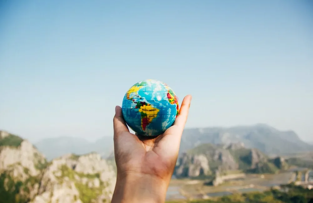 Hand holding a small globe with a blurry mountainous landscape in the background.