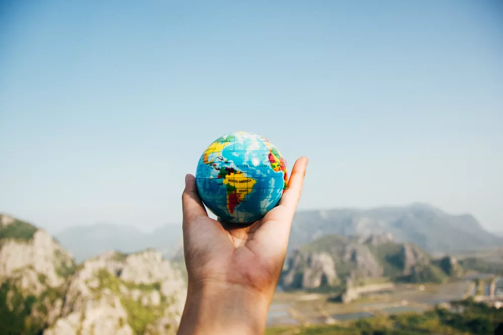 Hand holding a small globe with a blurry mountainous landscape in the background.