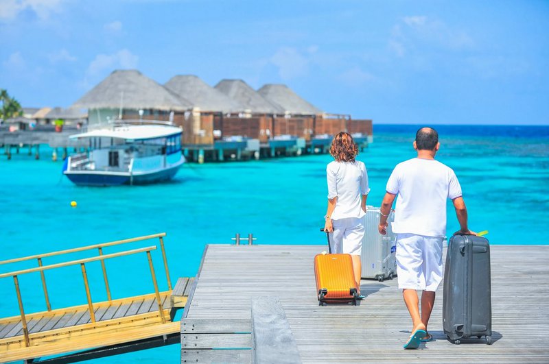 Two people with luggage walking on a jetty towards overwater bungalows with a boat nearby.