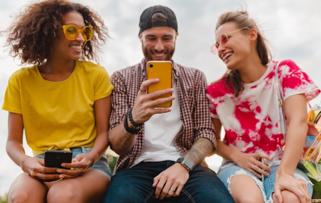 A group of three friends laughing and looking at a smartphone outdoors.