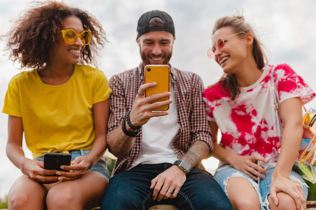 A group of three friends laughing and looking at a smartphone outdoors.