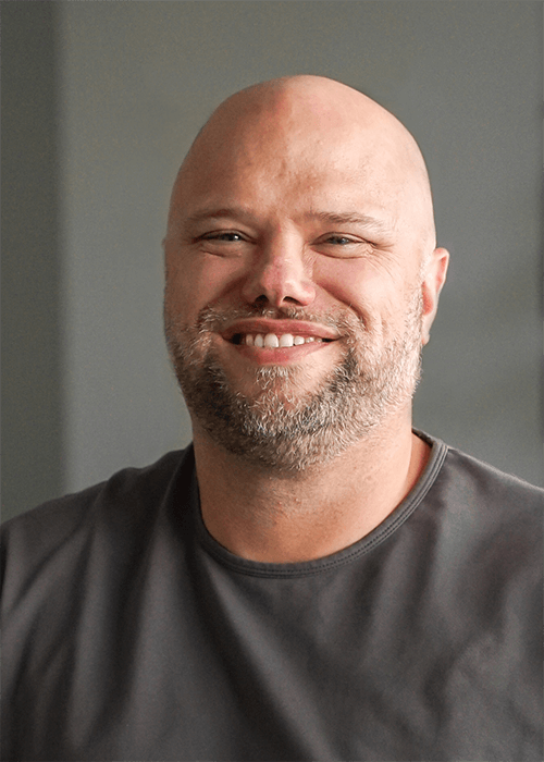 Portrait of a smiling man with a beard and a bald head wearing a grey shirt.