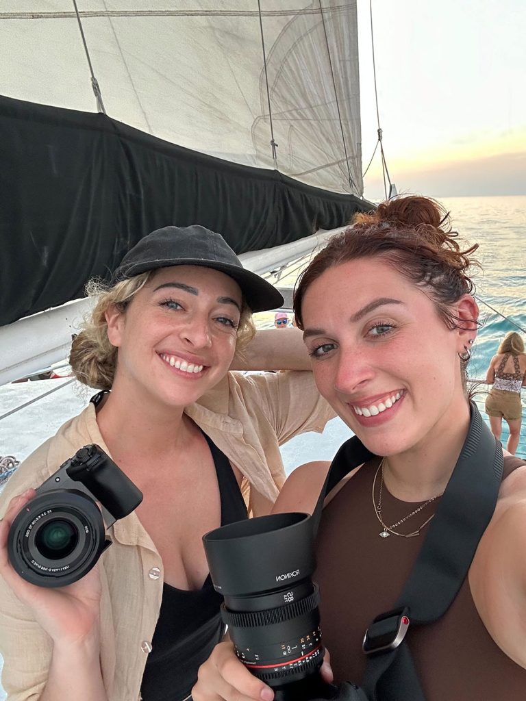 Two people holding cameras on a sailboat at sunset.